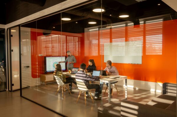 Group having a meeting in an orange room behind glass walls. A man giving a presentation in front of a long table.