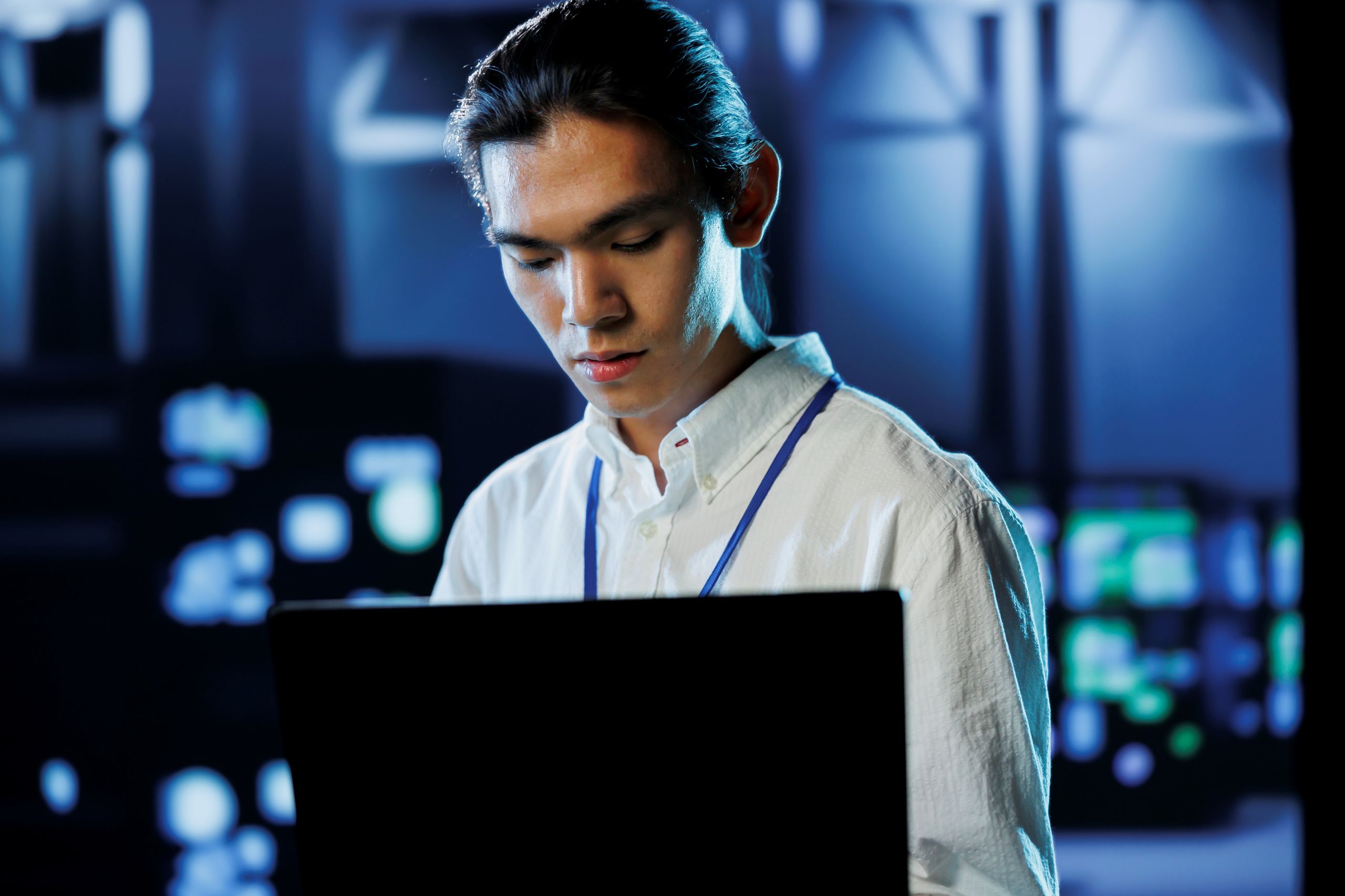 IT professional working on a laptop in a dimly lit server room with illuminated equipment in the background. IT professional working on a laptop in a dimly lit server room with illuminated equipment in the background